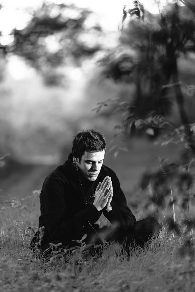 A man sitting in a grassy field, meditating with hands in prayer position, captured in black and white.