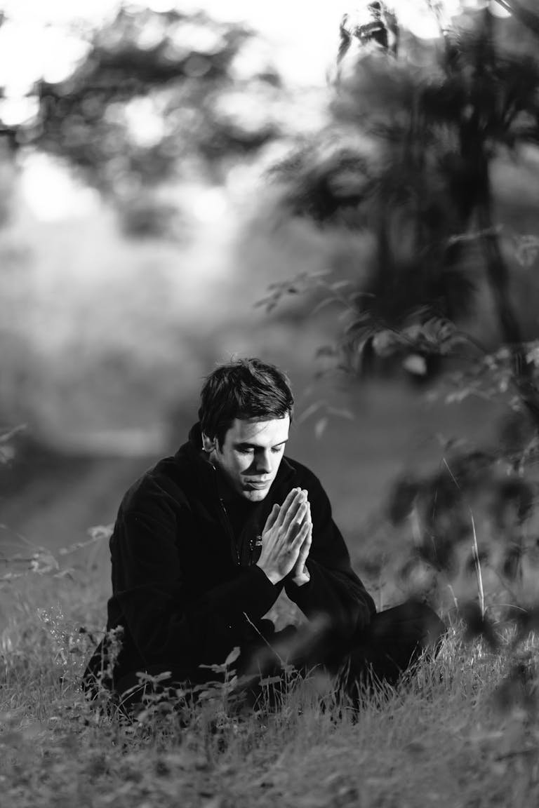 A man sitting in a grassy field, meditating with hands in prayer position, captured in black and white.