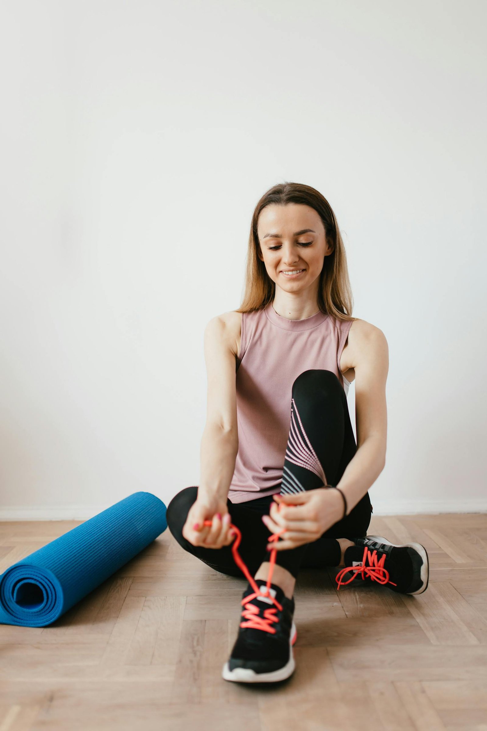 Full body positive female athlete in sportswear sitting on floor near folded blue fitness mat and tying sneakers before exercising indoors while looking down with smile